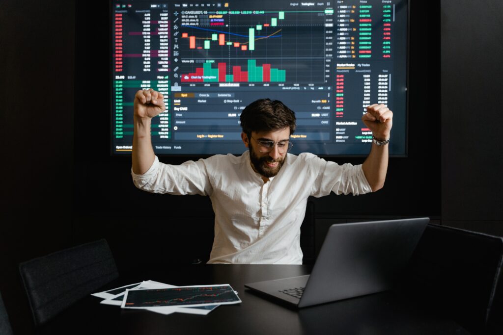 a photo of a man raising his 2 hands high because he just hit the jackpot in investing either passively or actively with graphs on the screen background