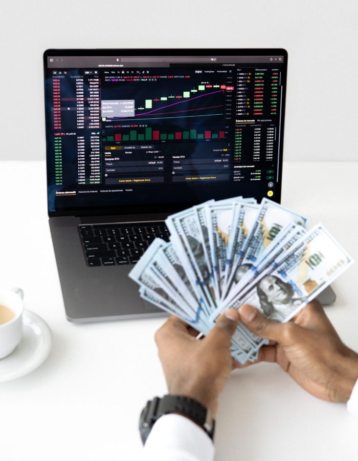 a picture of a hand handling a lot of dollar bills with a laptop on a background showing uptrend graphs that can be a result of a working private equity investment